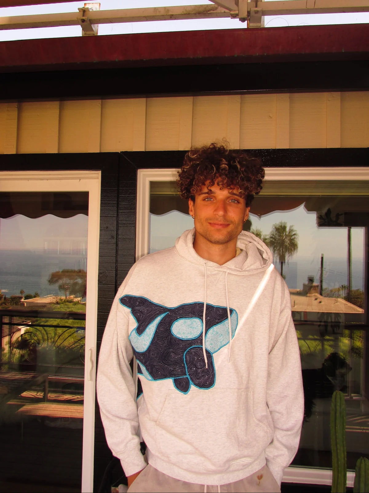 Ash orca hoodie worn on a sunny porch, showing the navy and turquoise stitched artwork.