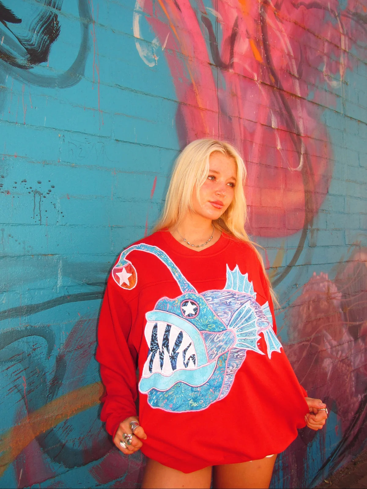Red anglerfish crewneck worn in front of a turquoise and pink mural, showing the full sewn fish.