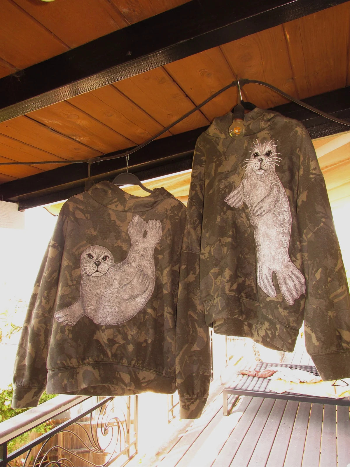 Curled and upright seal hoodies in camouflage hanging together under a wooden porch roof.