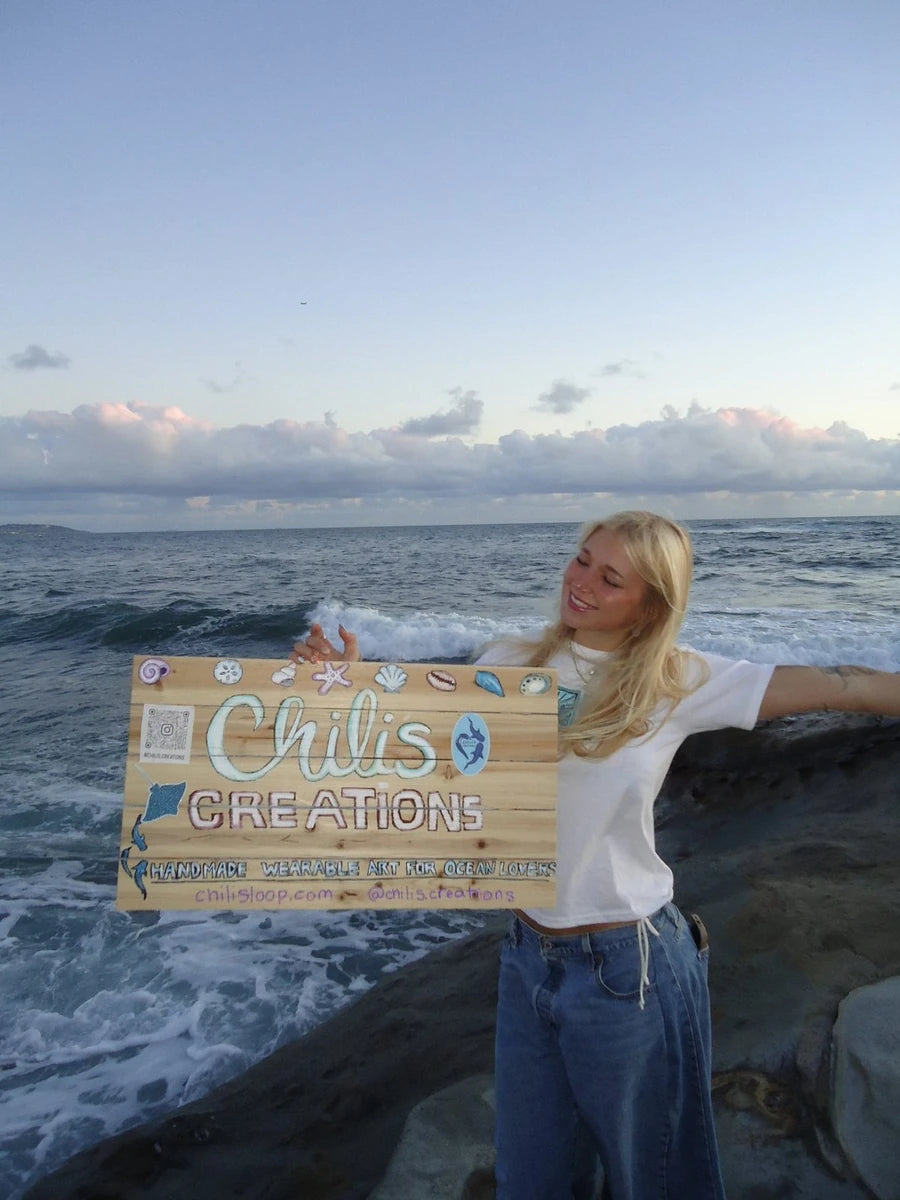 Chili Sloop holding her Chili’s Creations sign beside the ocean
